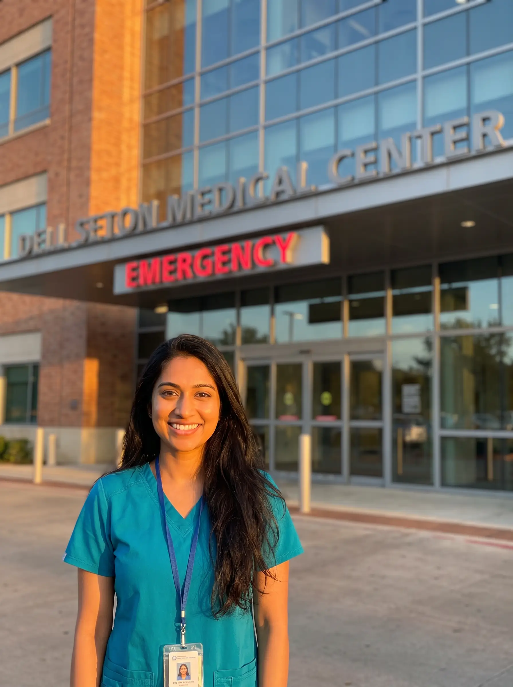 Neha in teal scrubs outside a hospital emergency entrance at sunset